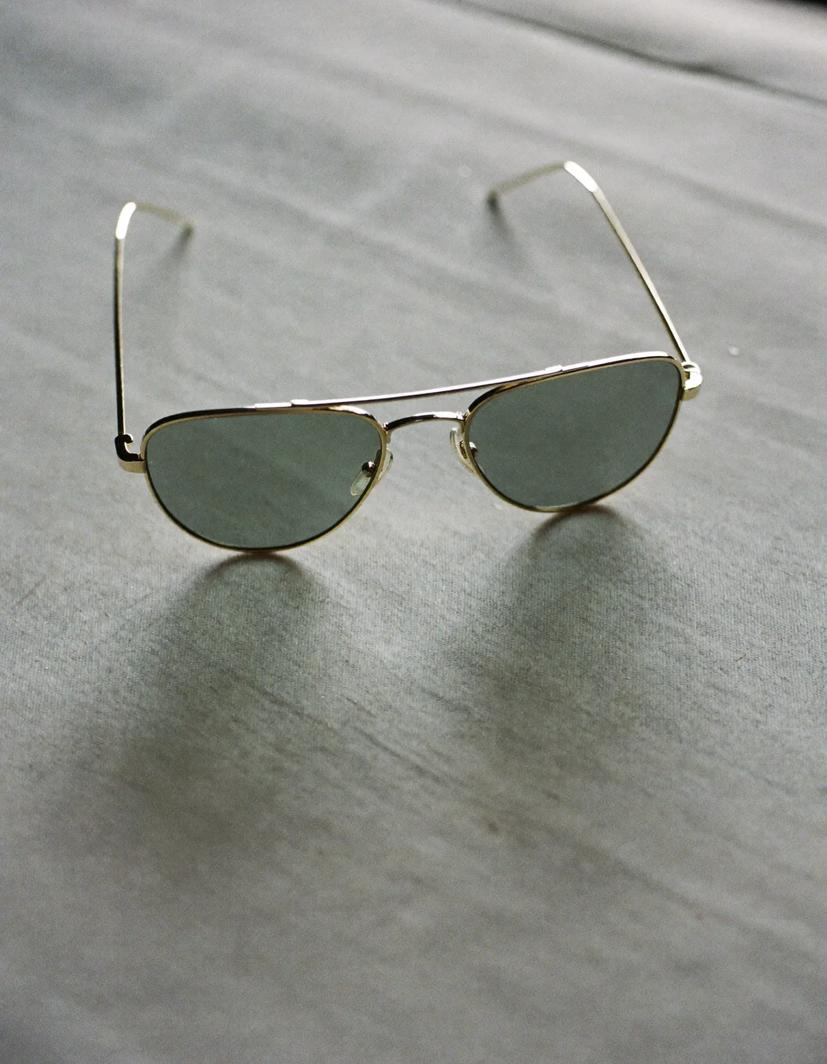 A pair of thin gold-framed aviator sunglasses with grey-green lenses lying open on pale linen