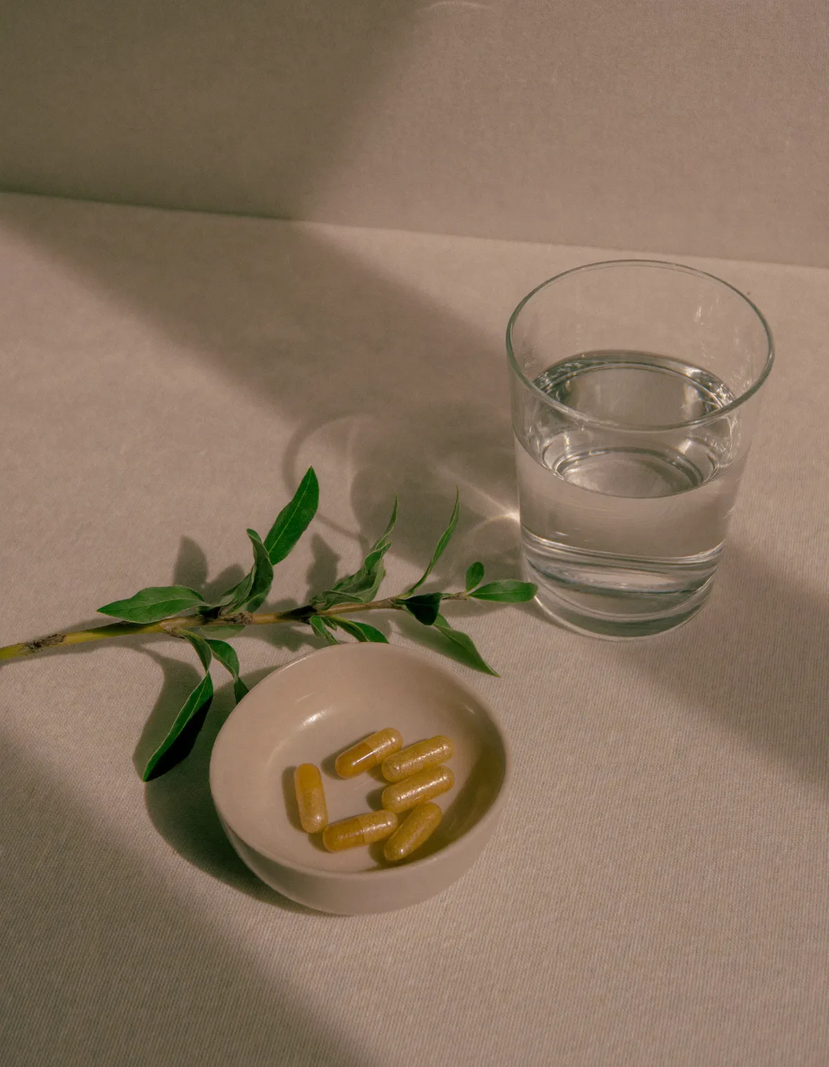 A few golden-yellow supplement capsules in a small cream ceramic dish beside a glass of water and green leaves