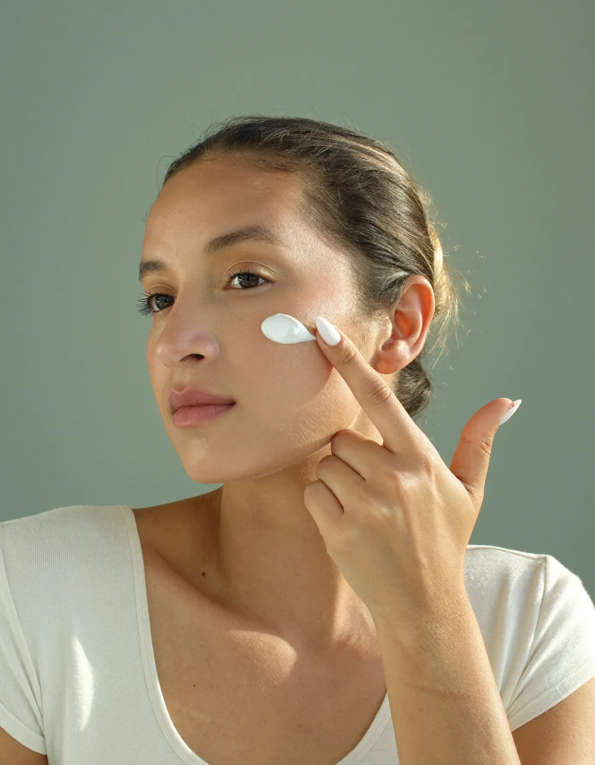 A woman with glowing skin applying a small dot of white sunscreen to her cheek, in a cream knit top