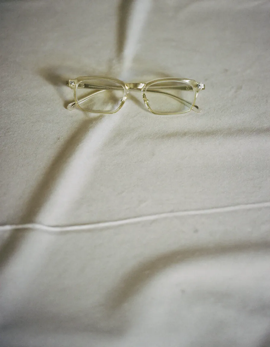 A pair of fine clear-framed eyeglasses folded on pale oyster linen in soft daylight