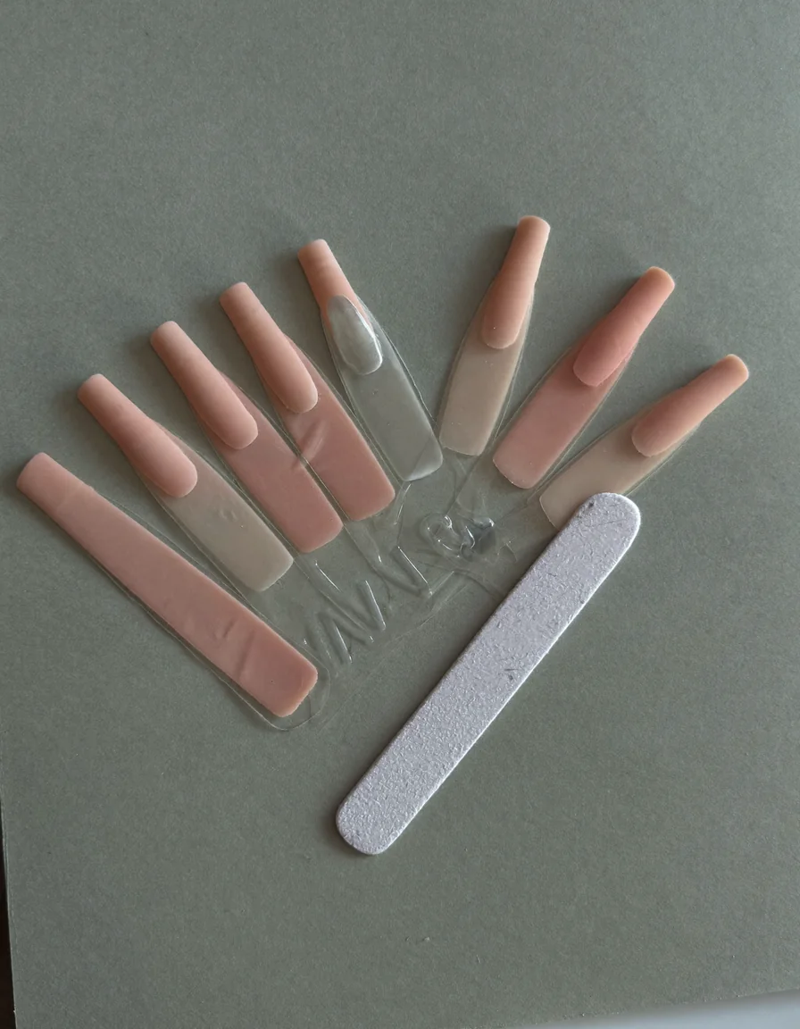 A fan of nude and soft rose nail tips beside a nail file on a pale oyster surface