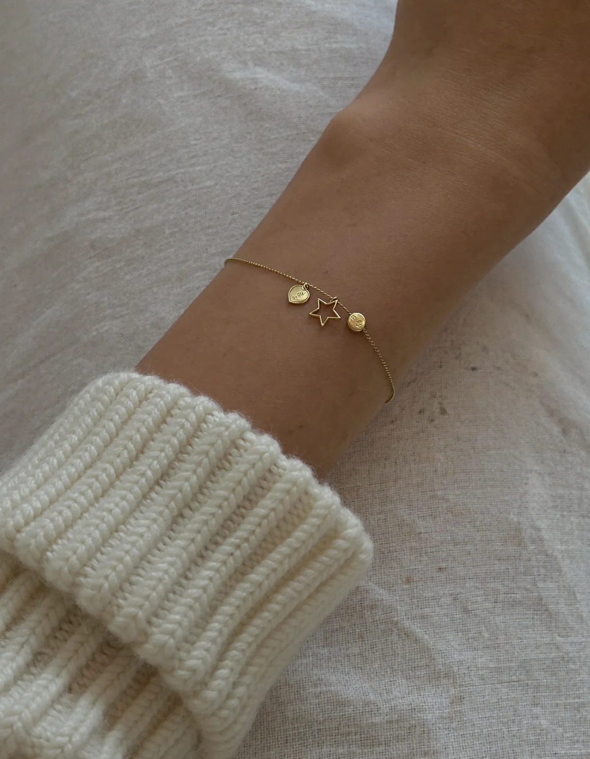 A slim wrist on oyster linen wearing a dainty gold charm bracelet with a tiny heart, star and disc