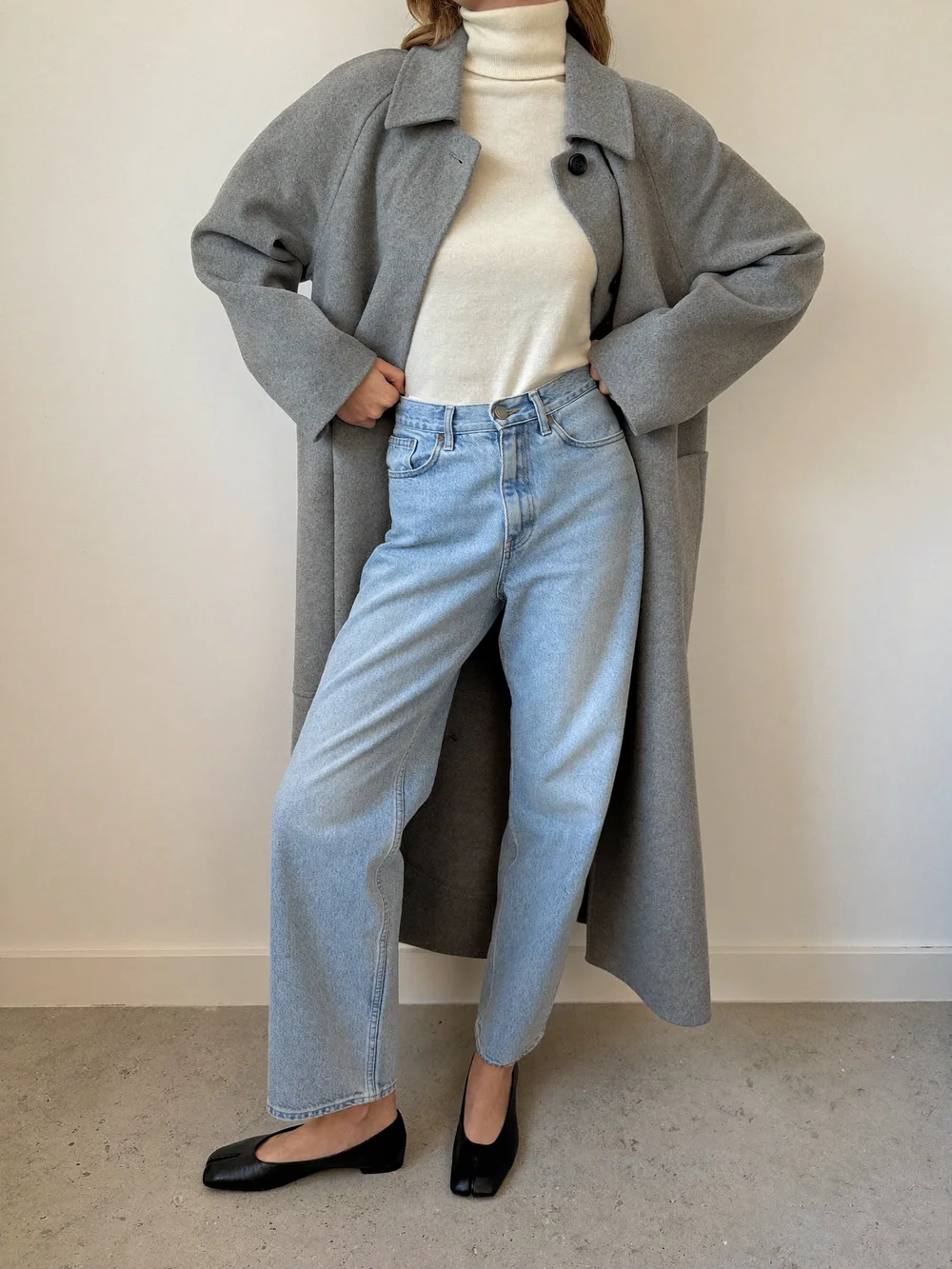 A woman against a cream wall in a long soft gray wool coat open over a cream turtleneck and straight jeans