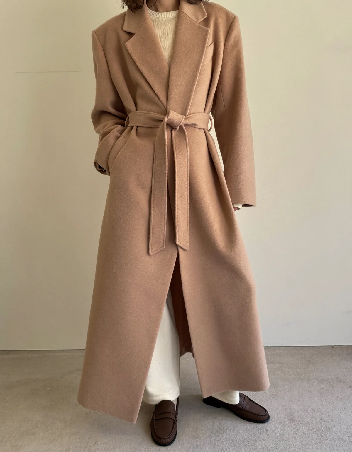A woman against a cream wall in a long camel wool wrap coat belted over a cream knit and straight trousers