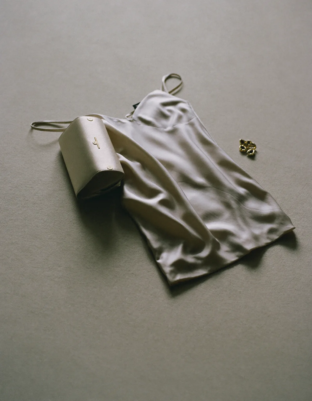 A champagne satin slip dress folded beside a small gold clutch and drop earrings on pale oyster linen