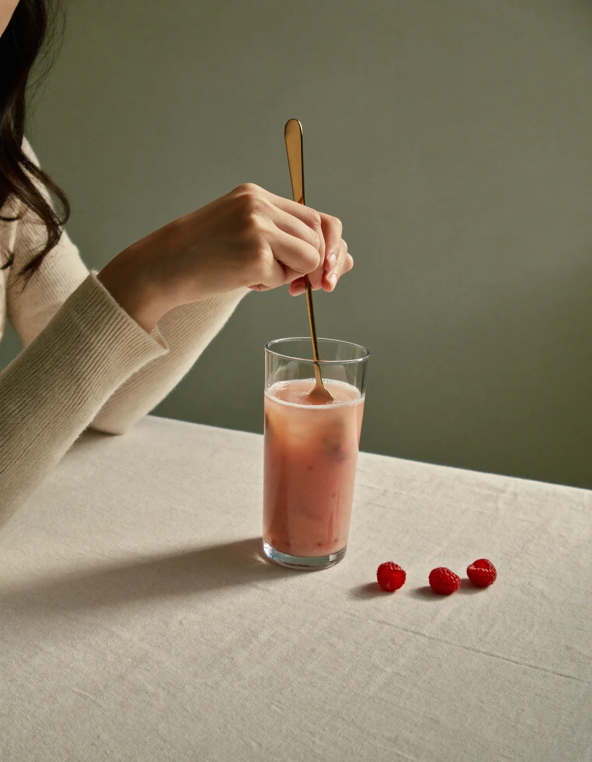 Hands in a cream knit sleeve stirring a pink berry drink with a gold spoon, raspberries beside it