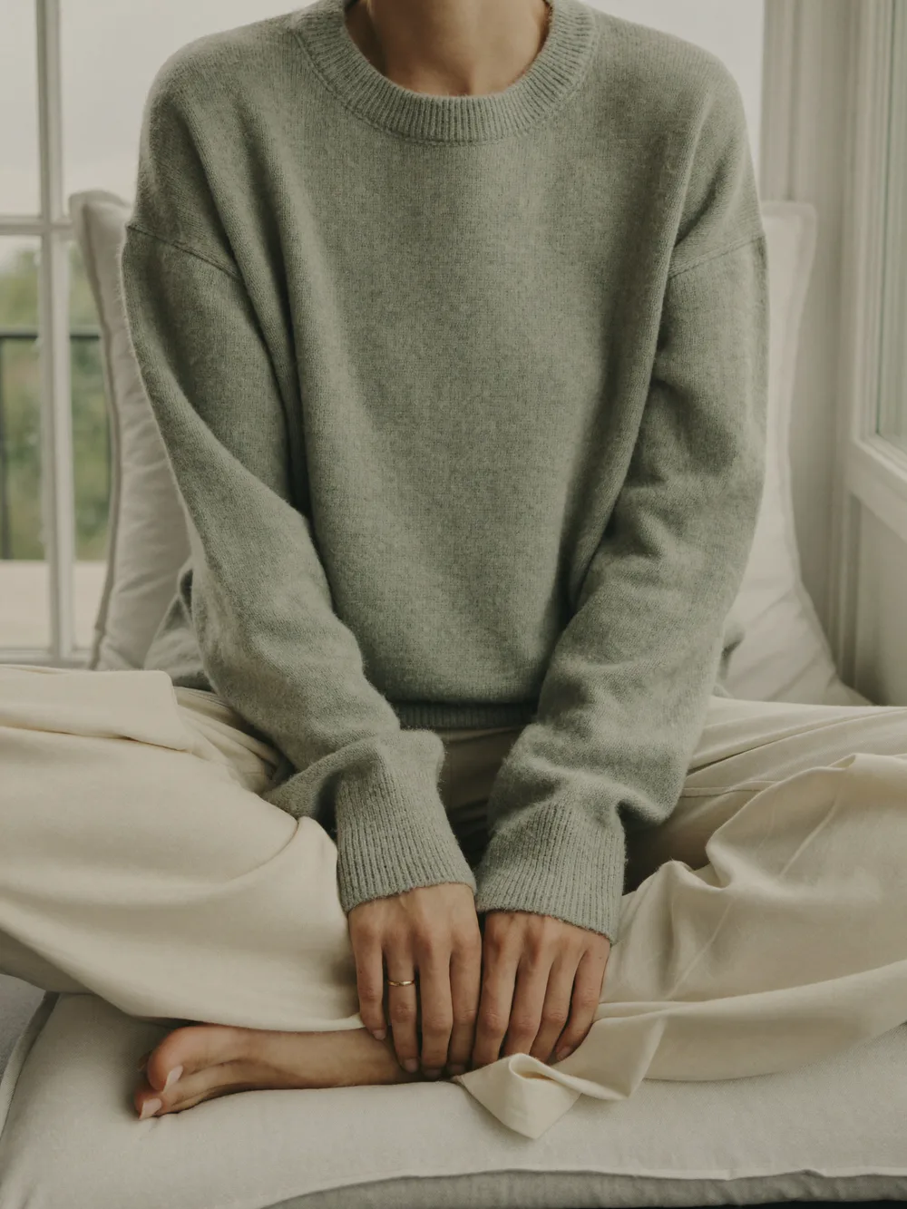 A woman in a long sage sweater and cream linen trousers sitting cross-legged on a cushion by a window, hands resting on her knees