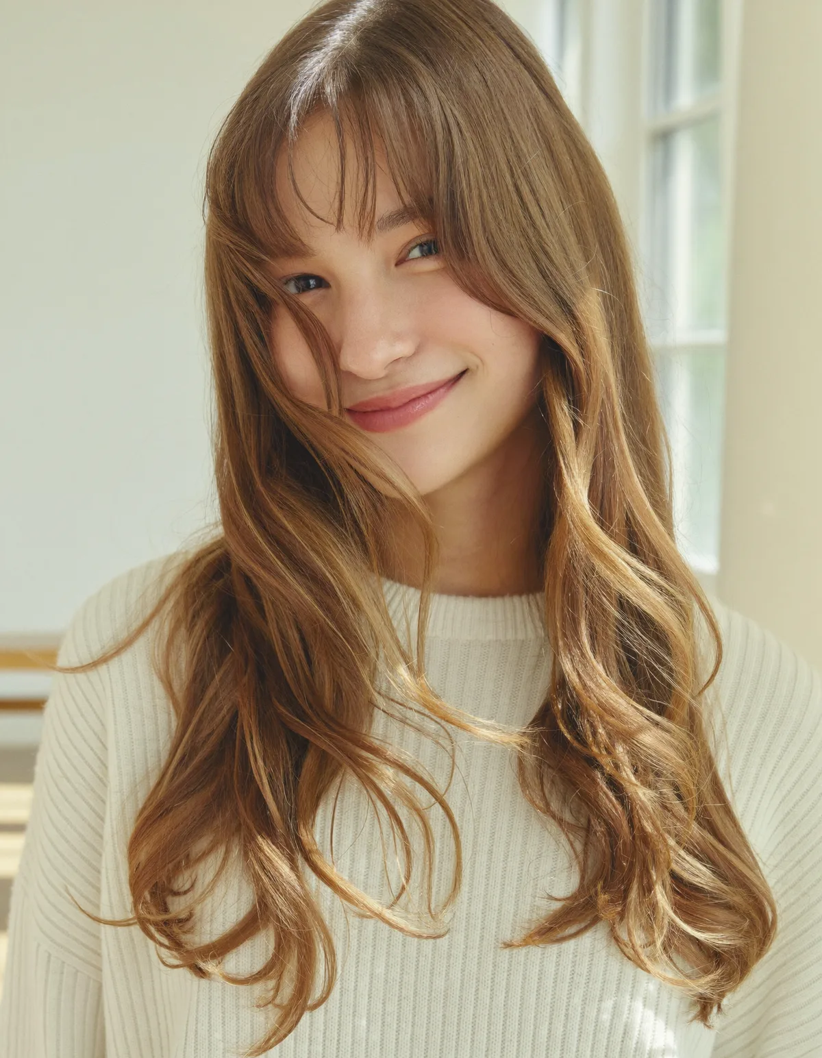 A smiling woman with long glossy light brown hair and soft center-parted curtain bangs, in a cream ribbed knit
