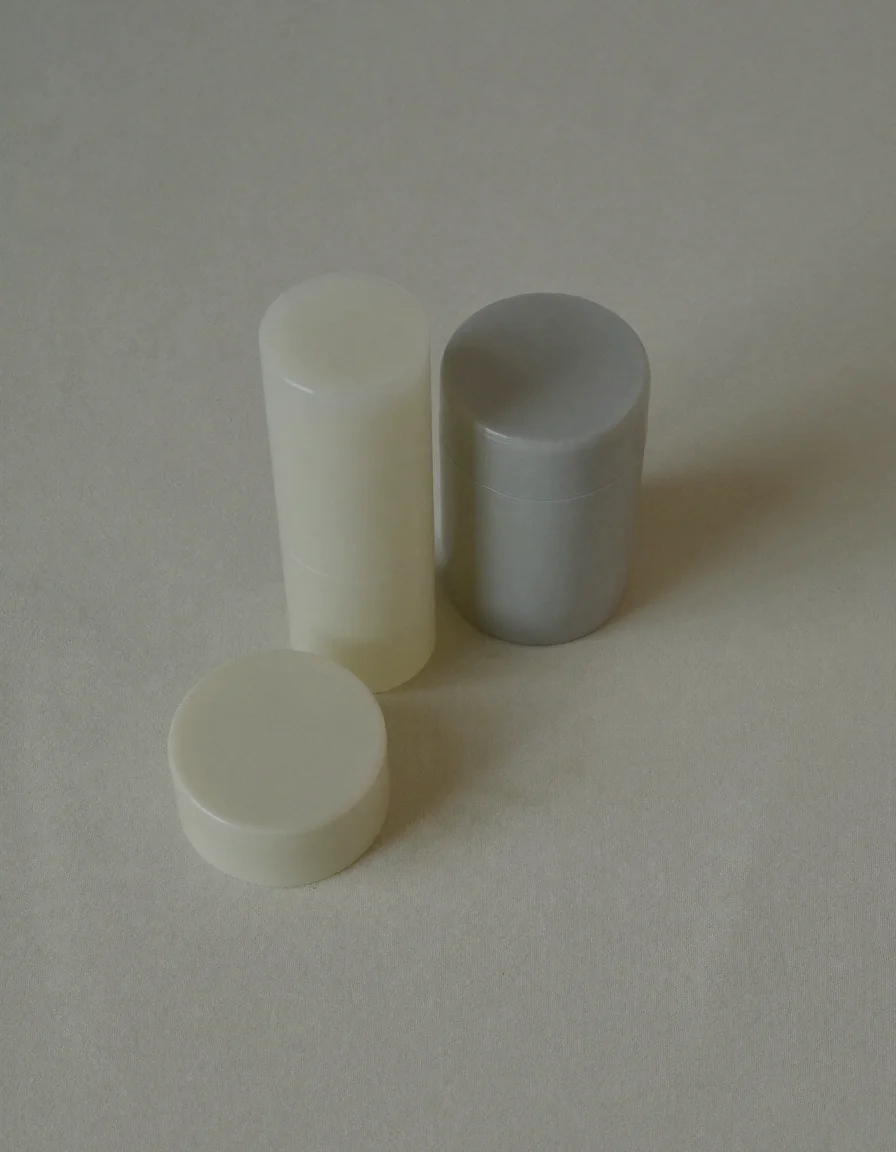 Two plain unlabelled cosmetic sticks, one ivory and one soft grey, on pale oyster linen