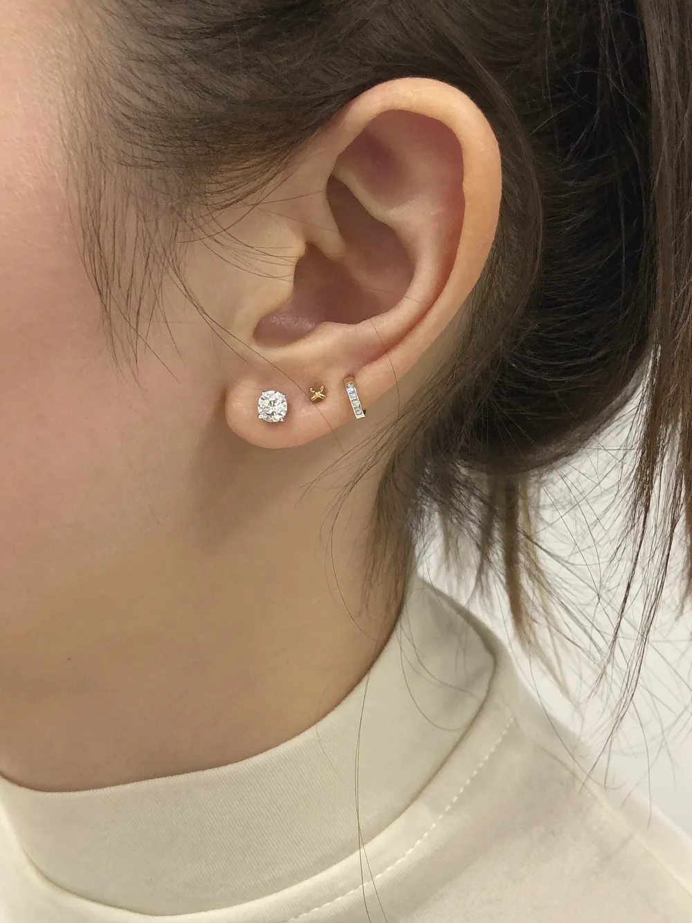 Close-up of an ear with a round diamond stud in the lobe and a tiny gold huggie in a second piercing