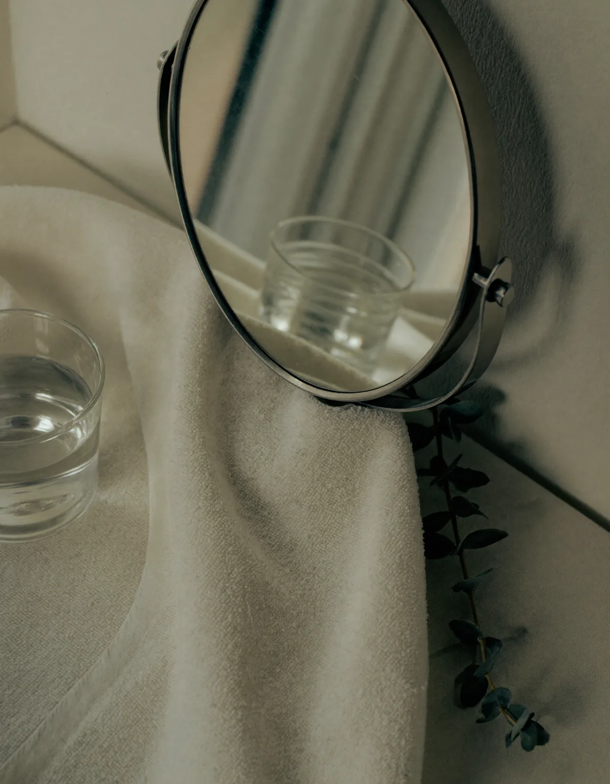 A small round vanity mirror leaning on a folded ivory towel beside a glass of water and a eucalyptus sprig