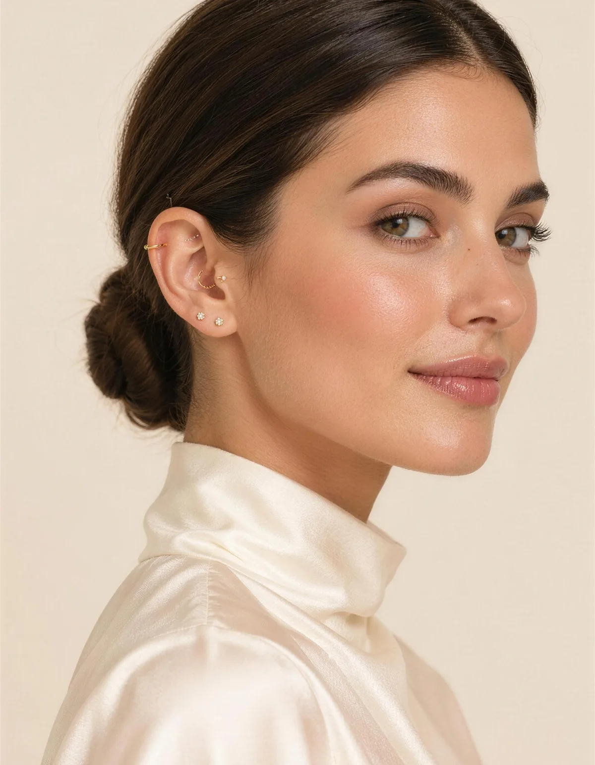 A woman in three-quarter profile with a sleek bun and a curated ear: lobe studs, a gold helix hoop, a conch ring and a tiny tragus stud