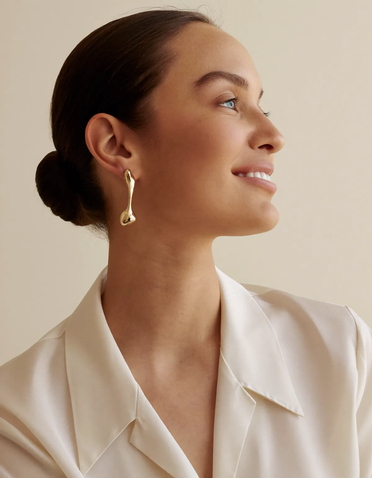 A smiling woman in profile with a sleek low bun wearing a sculptural gold drop earring and an ivory blouse