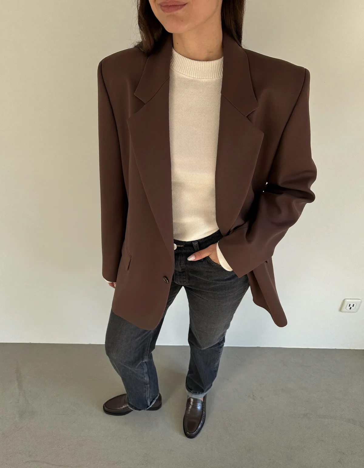 A woman against a cream wall in a structured chocolate brown blazer, cream knit, straight dark jeans and loafers