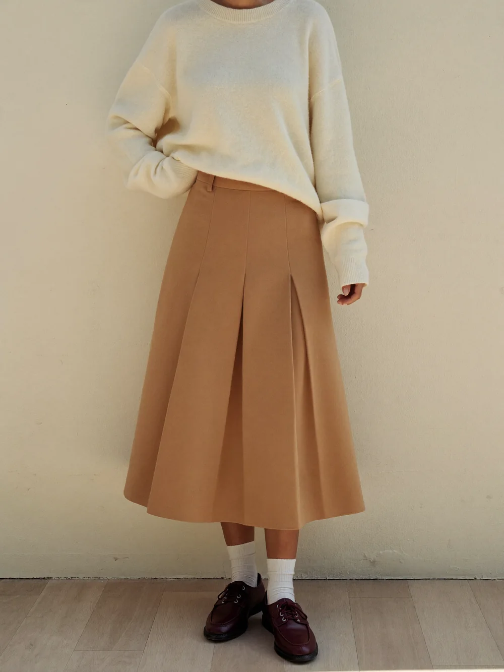 A woman against a cream wall in a cream sweater, camel pleated midi skirt, white socks and burgundy loafers