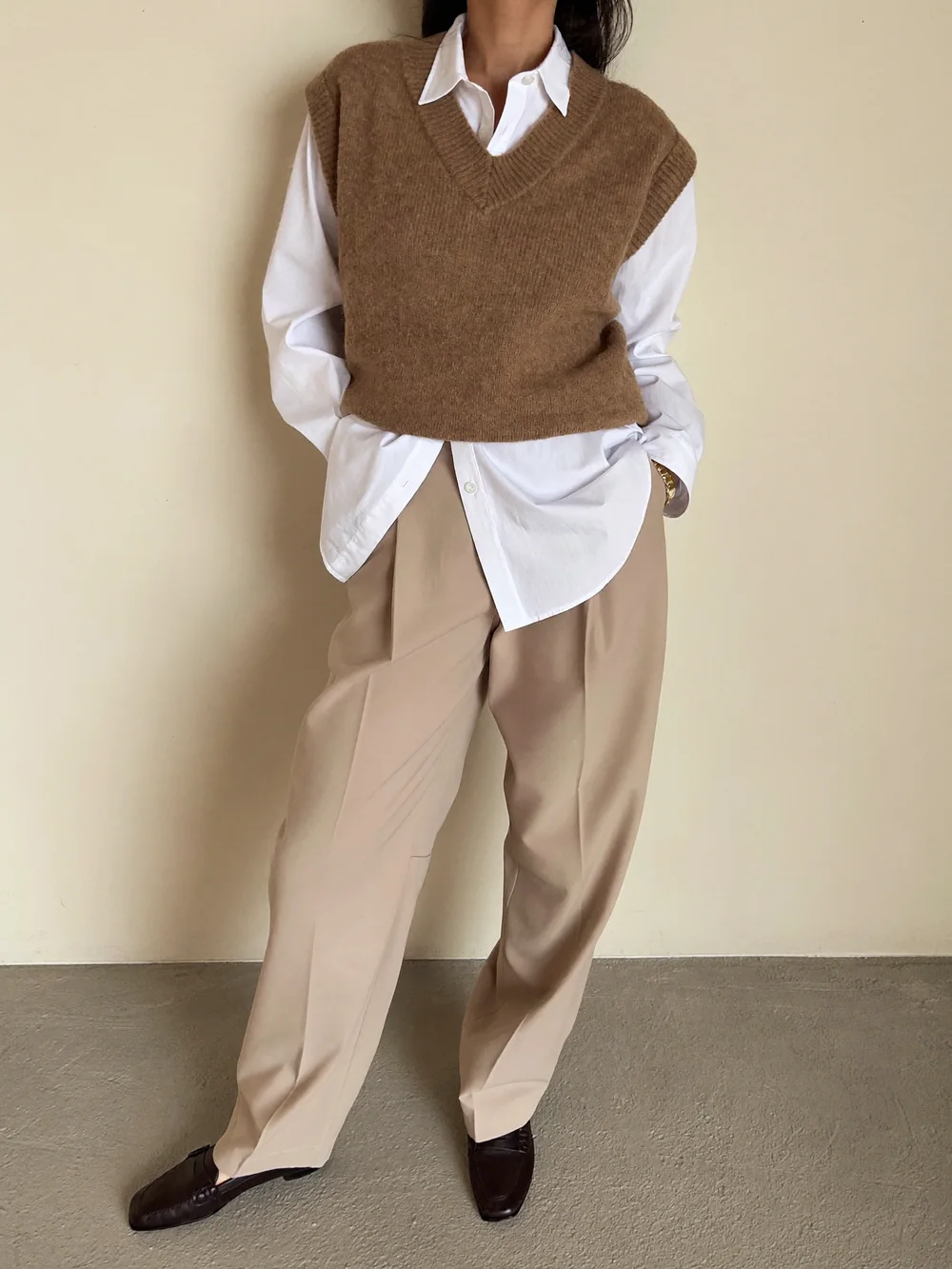 A woman against a cream wall in a brown knit vest over a white shirt, beige wide-leg trousers and brown loafers