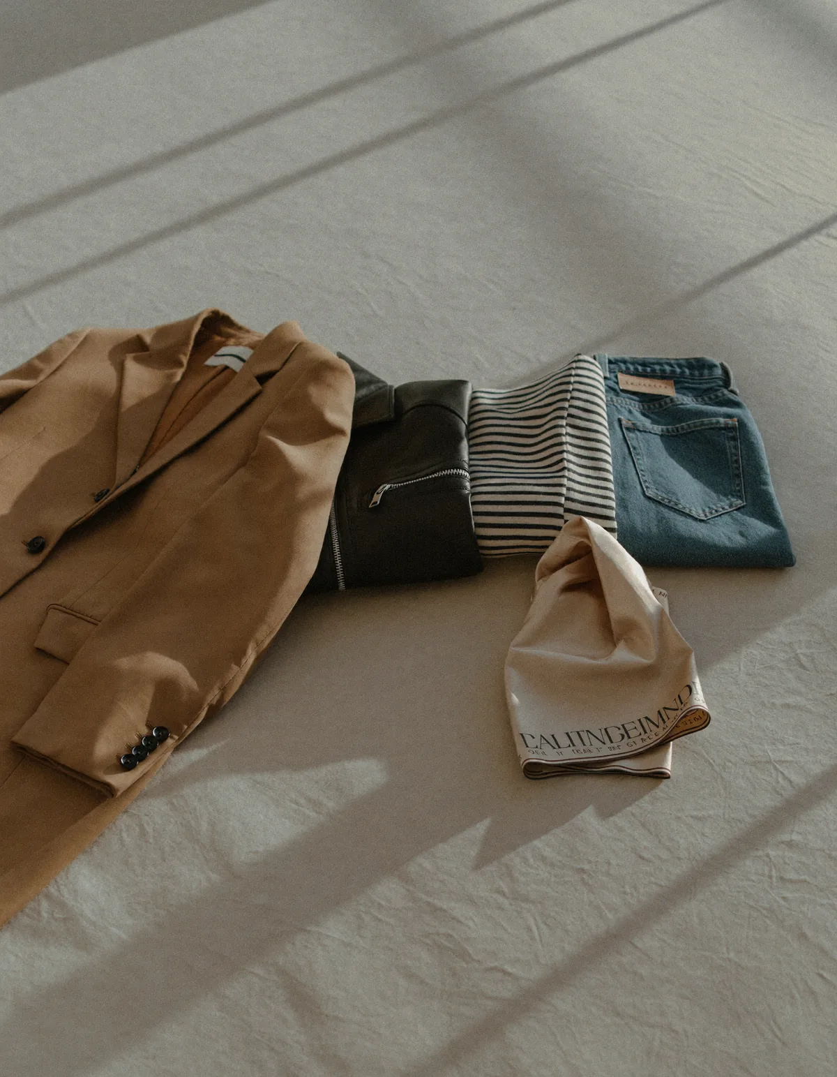 Five folded garments in different textures in a row: camel blazer, black leather jacket, striped knit, denim and a silk scarf