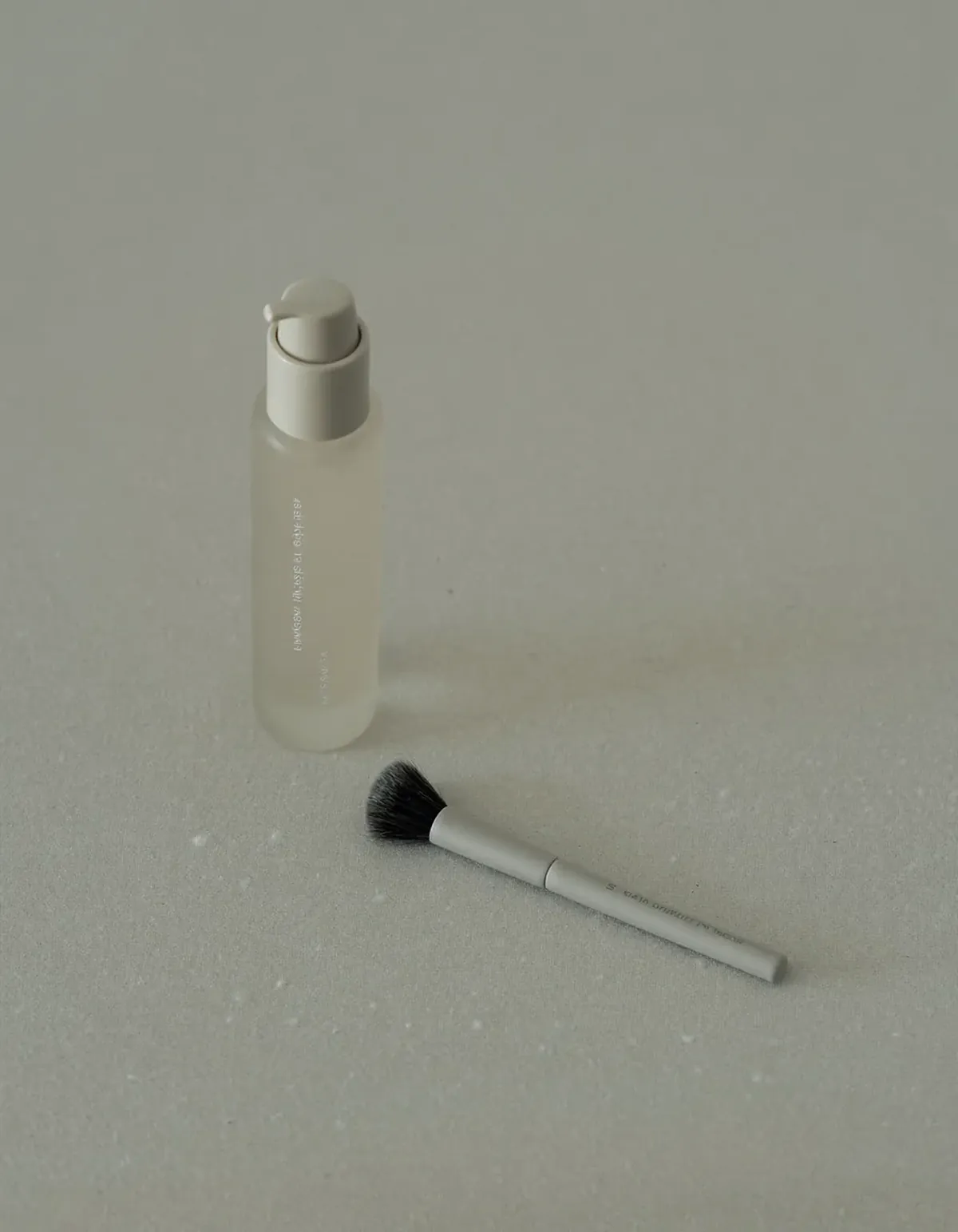 A slim frosted glass bottle with a pale pump top standing beside a small flat makeup brush