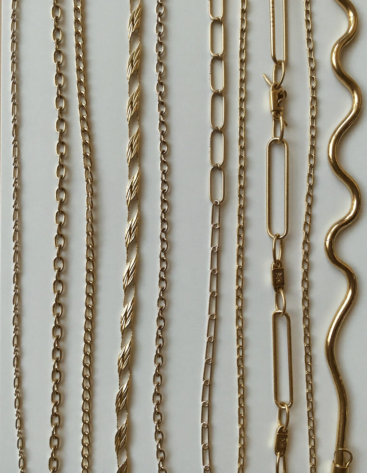 Six different gold necklace chains laid in parallel curves on pale oyster linen