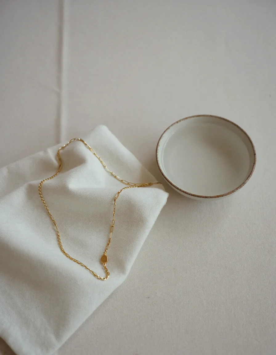 A fine gold chain on a folded white cloth beside a shallow pale bowl of clear water