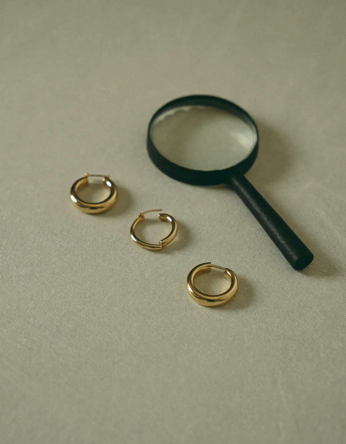 Three small gold hoop earrings of different thickness in a row beside a jeweler's loupe