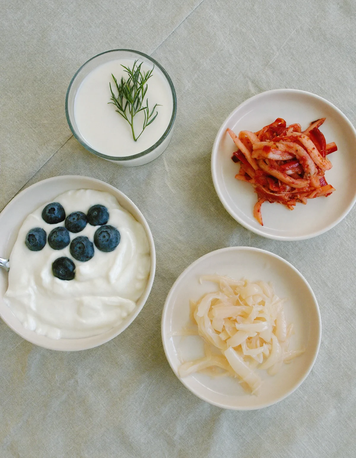 Bowls of yogurt with blueberries, kimchi and sauerkraut beside a glass of kefir on oyster linen