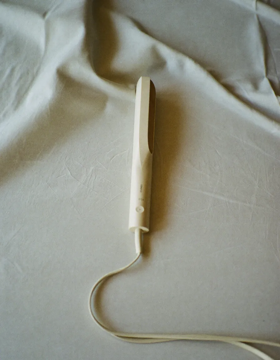 A slim cream-coloured styling wand lying diagonally on pale oyster linen