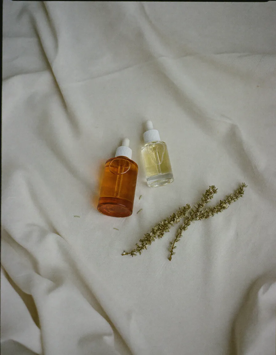 Two small amber glass dropper bottles and a sprig of dried herb on pale oyster linen