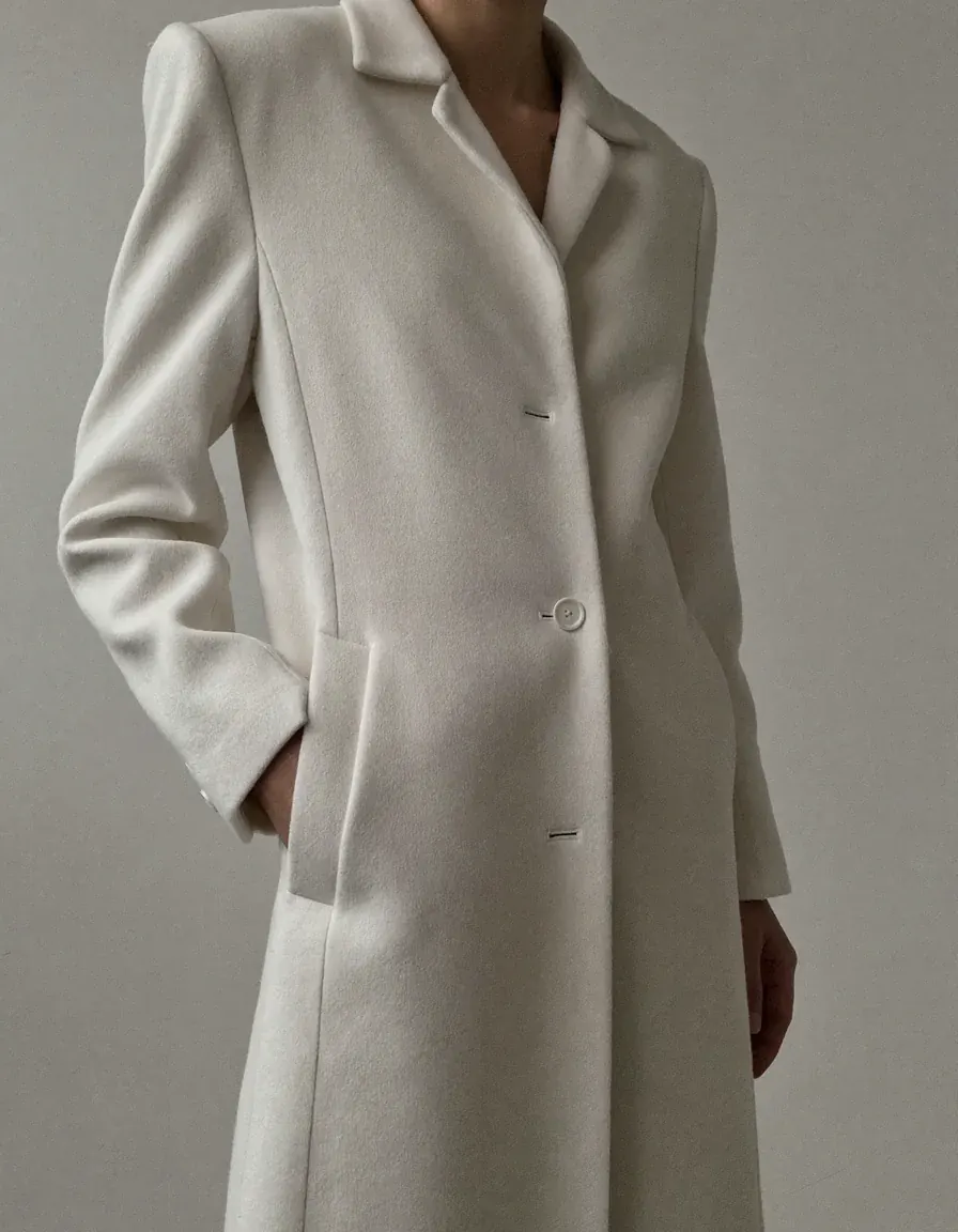 A woman wearing an impeccably tailored ivory wool coat, one hand resting in the pocket