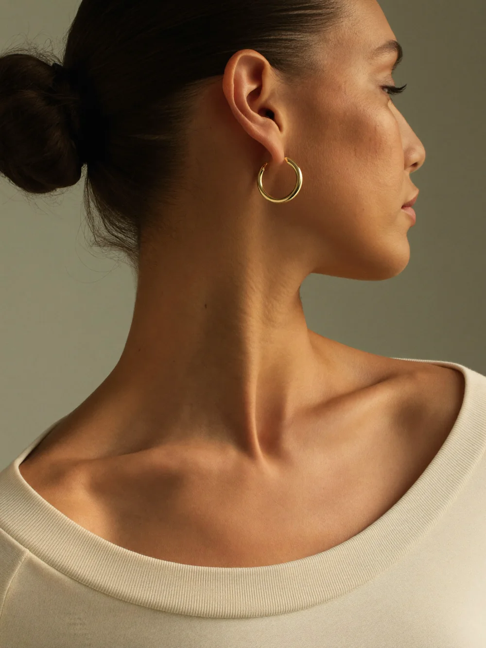 A woman in profile with a sleek low bun wearing a polished medium gold hoop earring and a cream top
