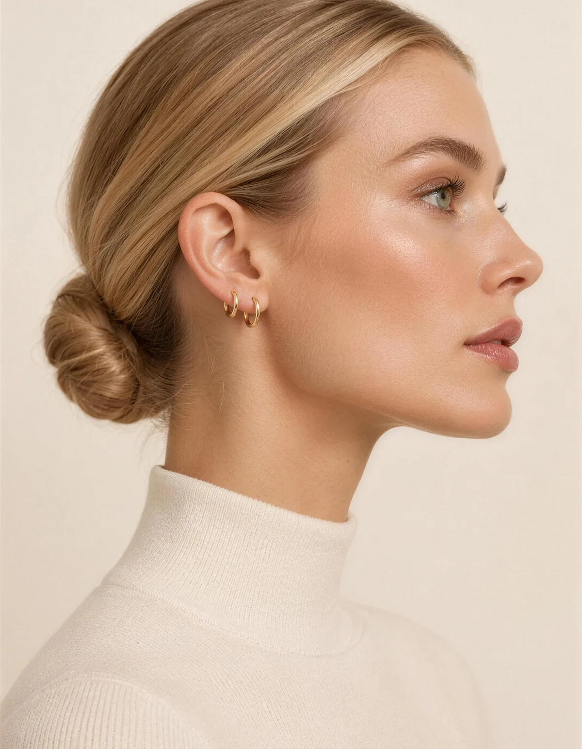 A woman in profile with a sleek honey-blonde bun wearing two thin gold huggie earrings that hug the lobe, in an ivory knit