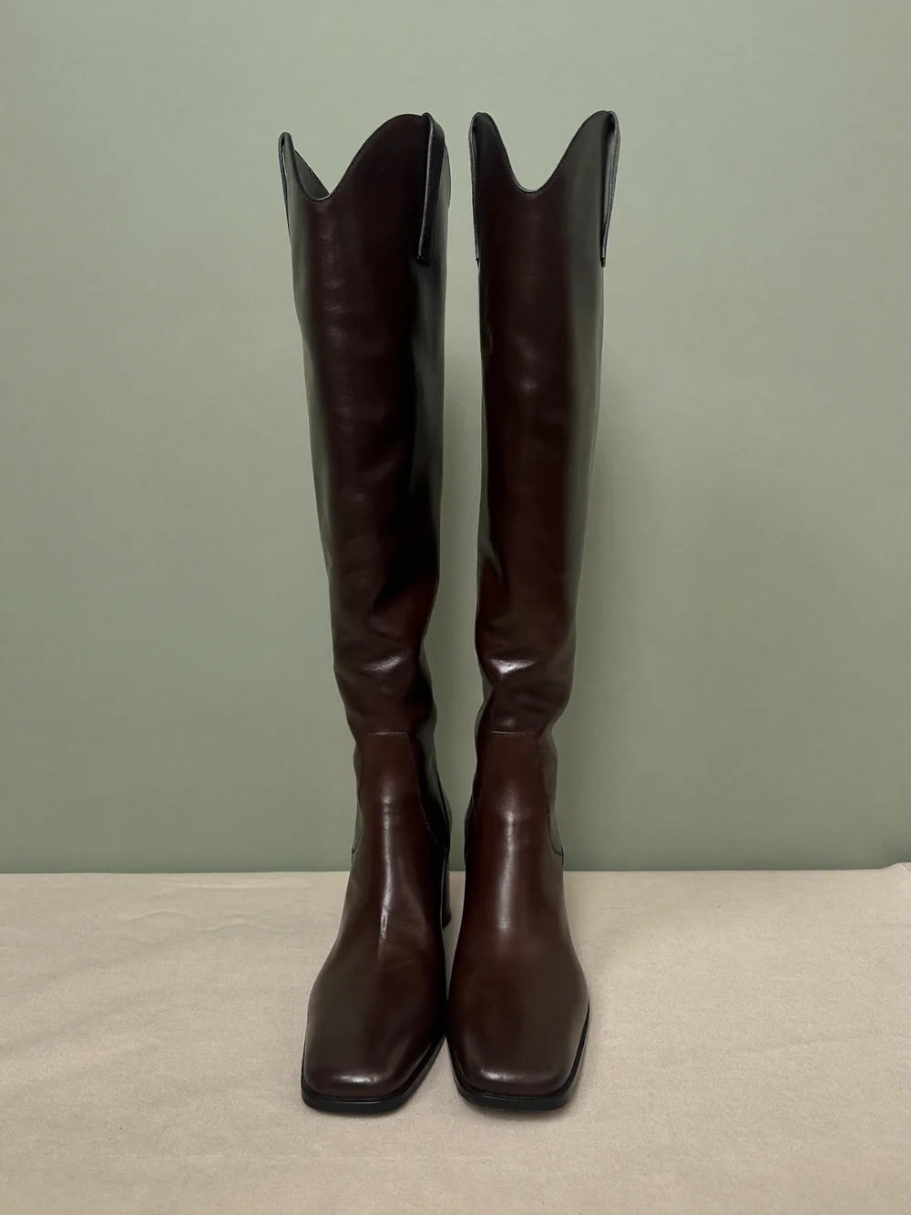 A pair of dark brown leather knee-high boots with scalloped western-style tops standing on oyster linen against a sage wall