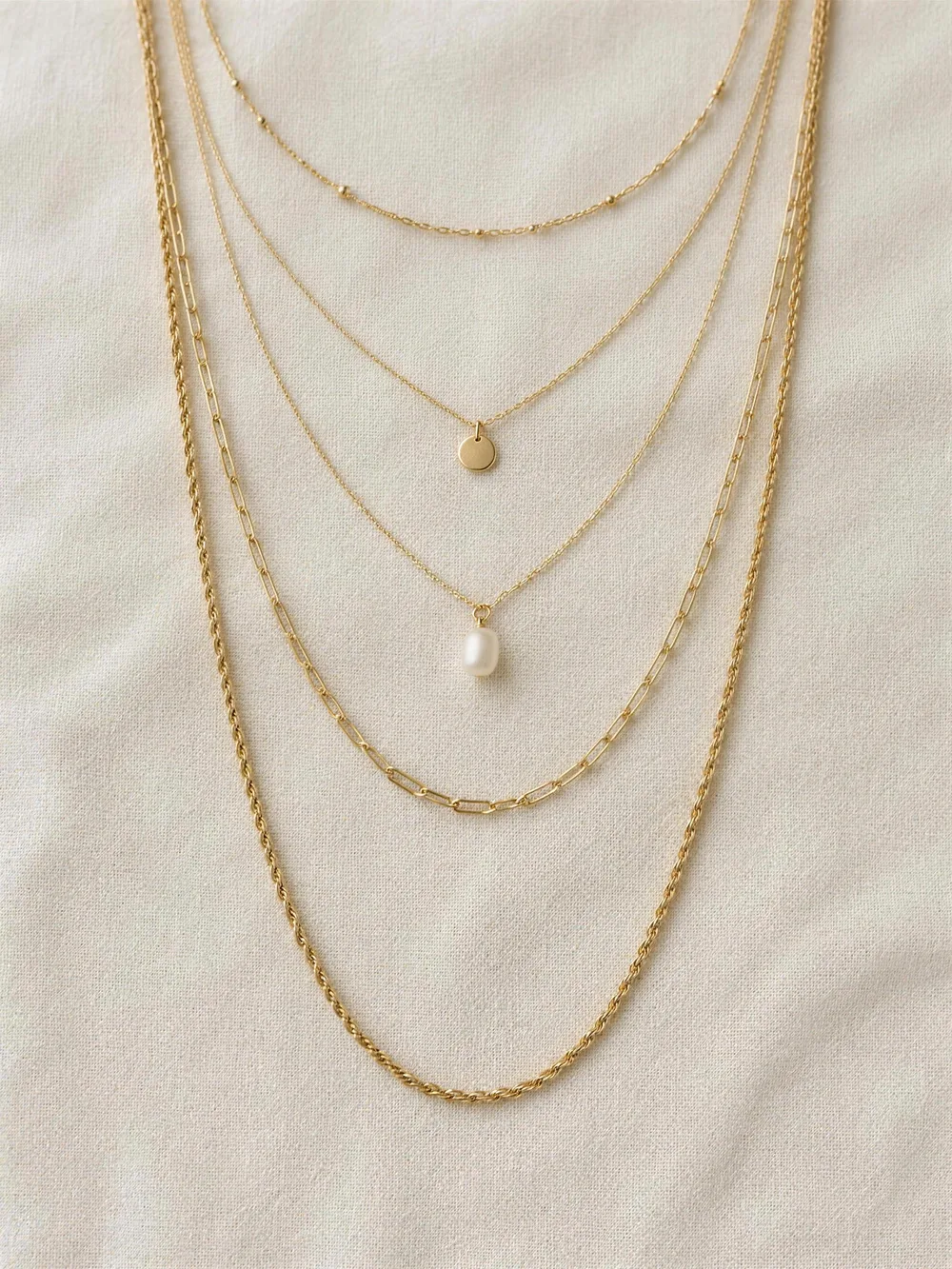 Five delicate gold necklaces on linen in graduated lengths: a fine choker, disc pendant, pearl pendant, paperclip and rope chain