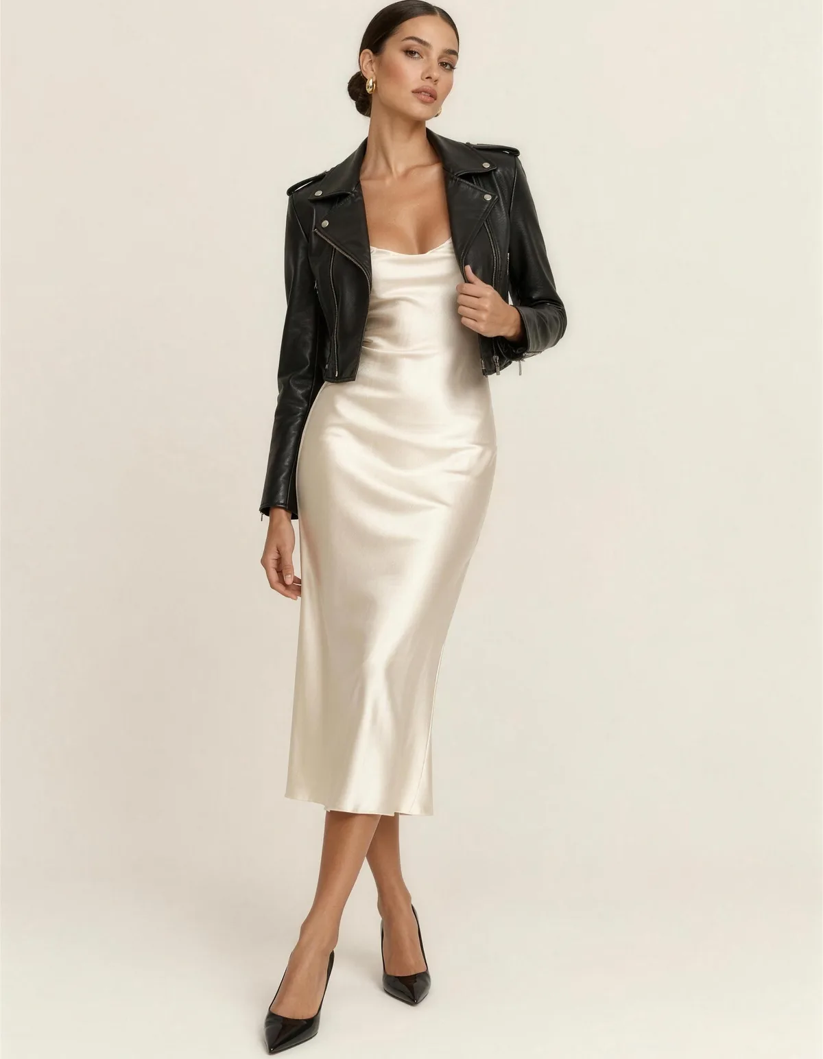 A beautiful woman in a cropped black leather moto jacket worn open over a cream satin slip midi dress with black pointed heels