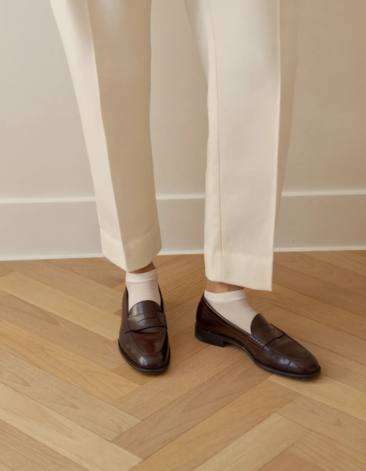 Feet on a pale oak floor in dark brown leather penny loafers with thin cream socks under cream cropped trousers