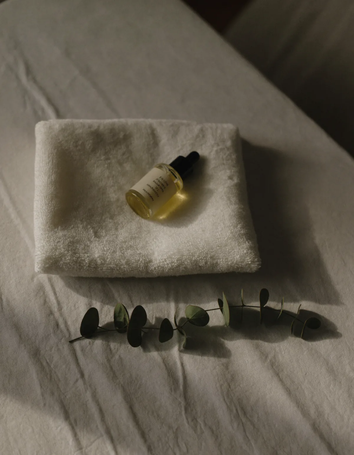 A folded white cotton towel, a small glass dropper bottle of face oil and a sprig of eucalyptus on pale oyster linen
