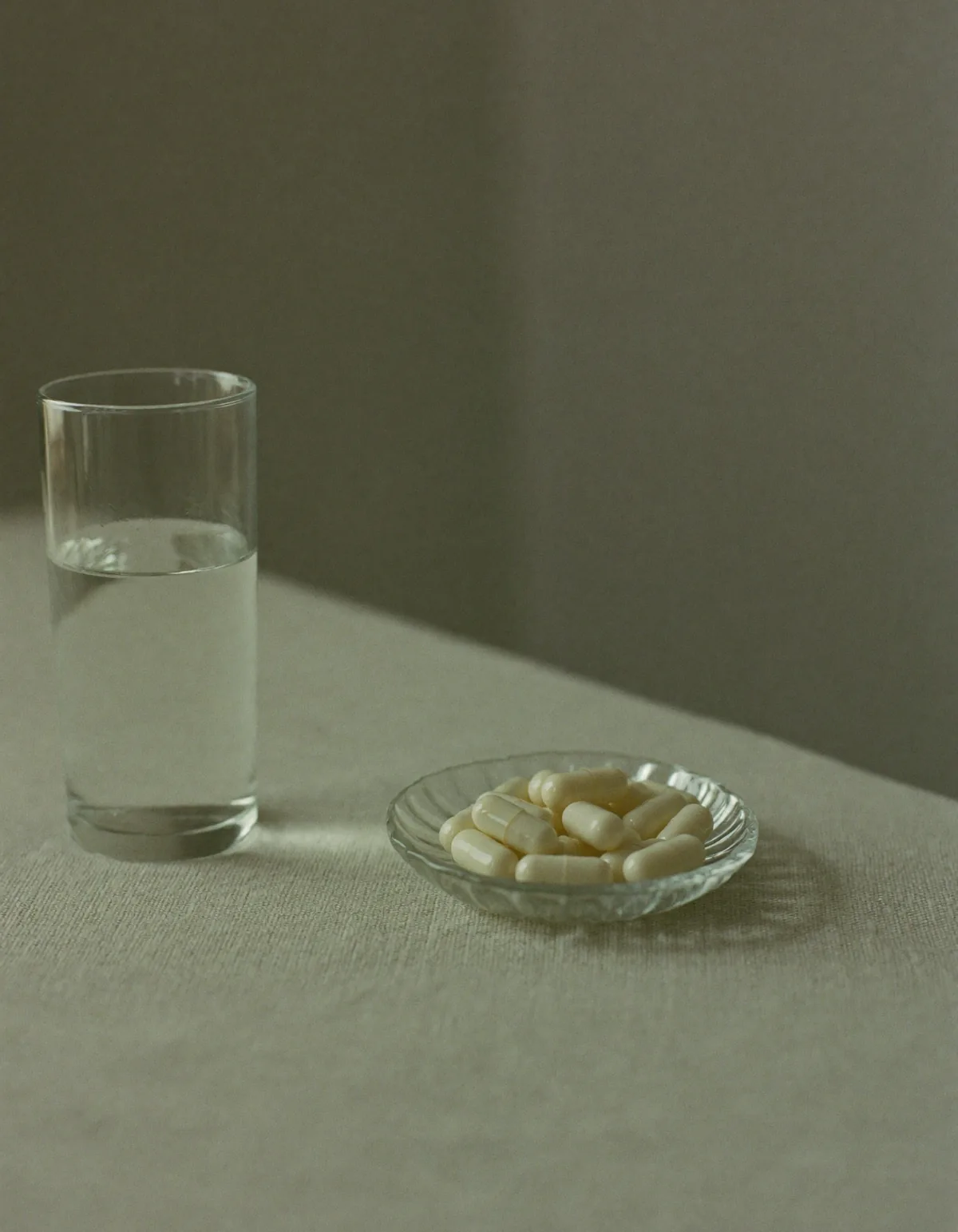 A small clear glass dish of cream coloured capsules beside a tall glass of water on pale oyster linen