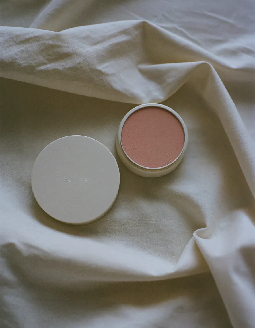 Three open cream and rose powder compacts beside a slim lipstick on pale oyster linen