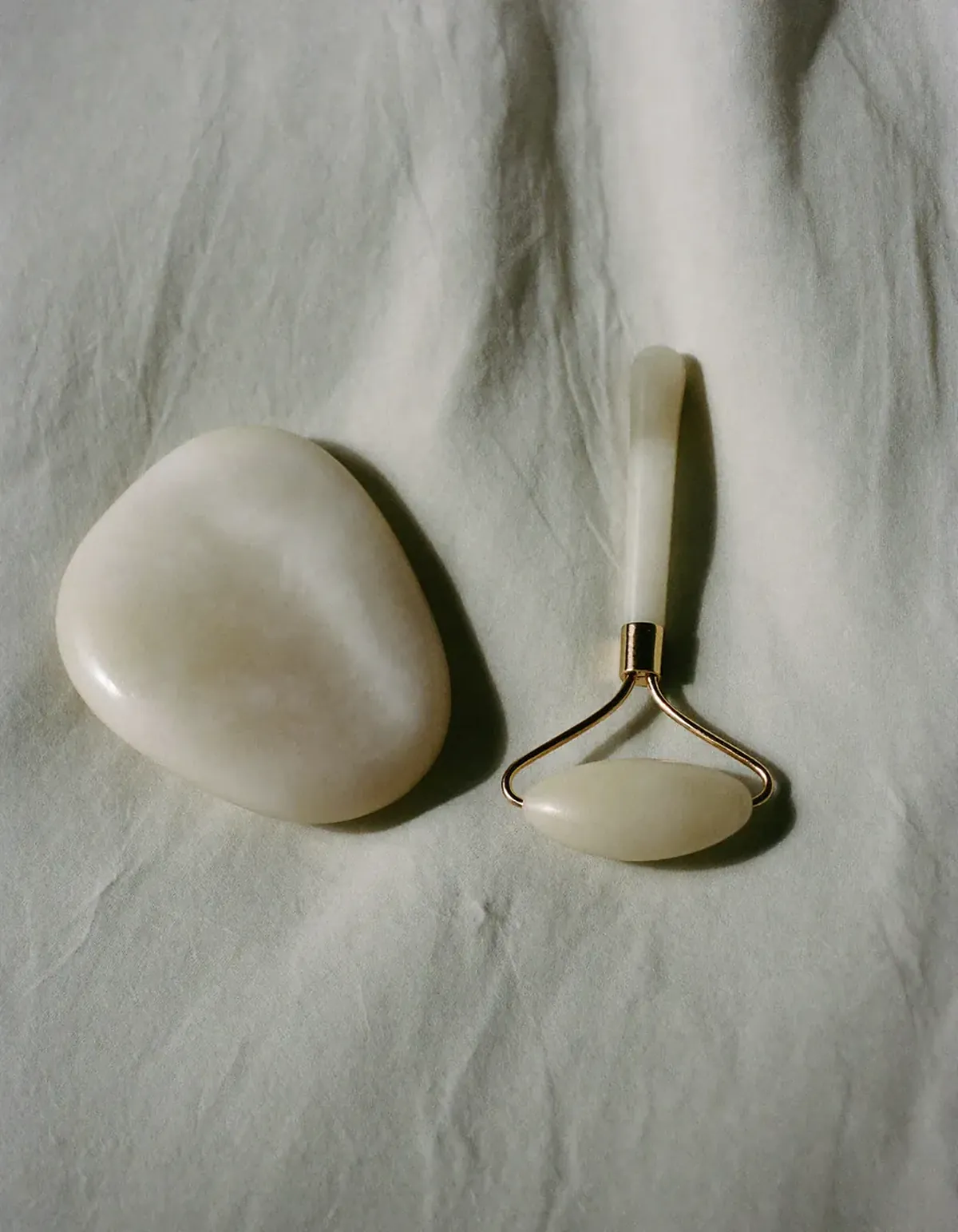 A pale jade gua sha stone beside a small rounded facial massage tool on pale oyster linen