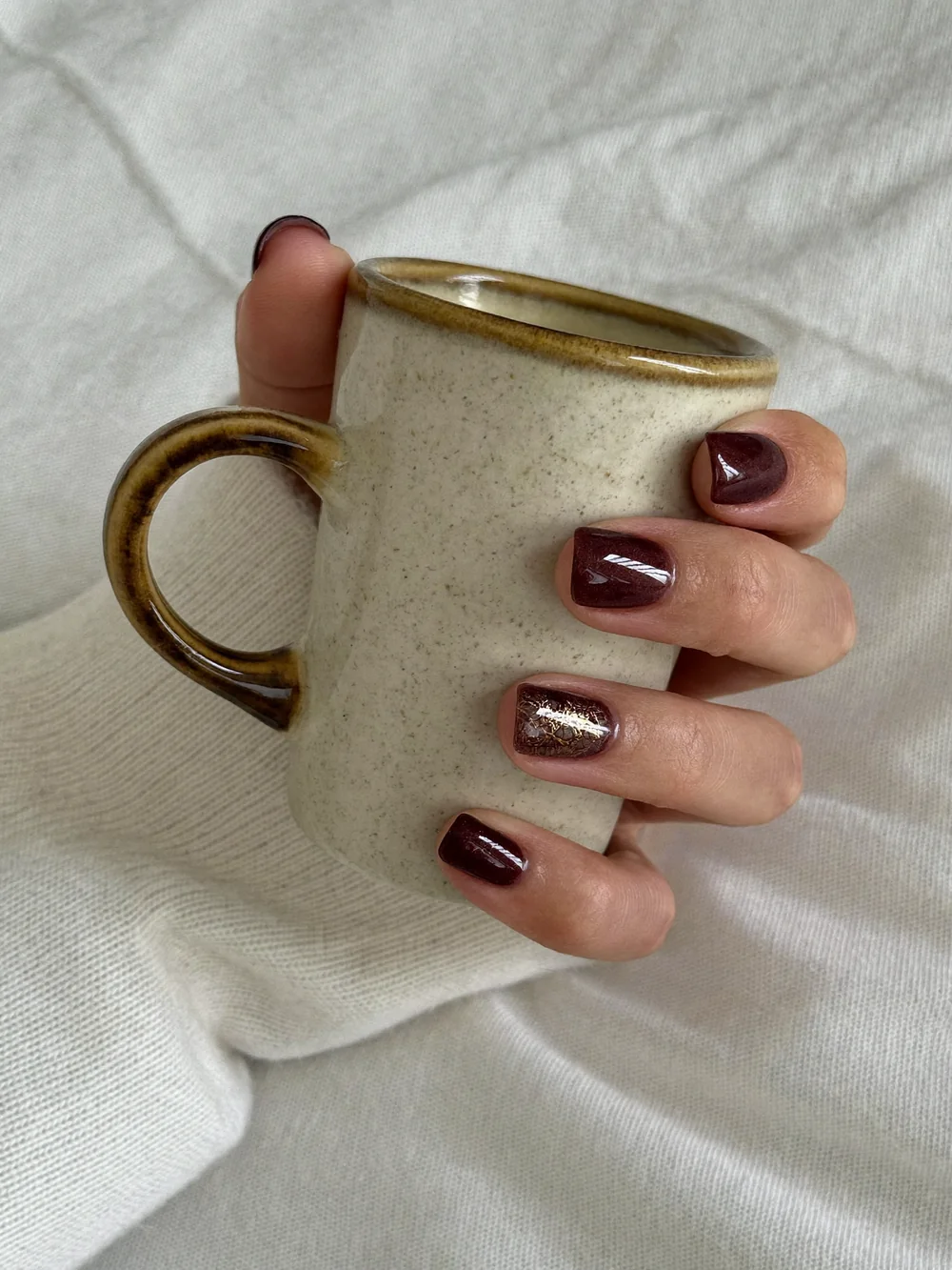 A hand holding a ceramic mug, with short burgundy cat eye nails and one glitter accent