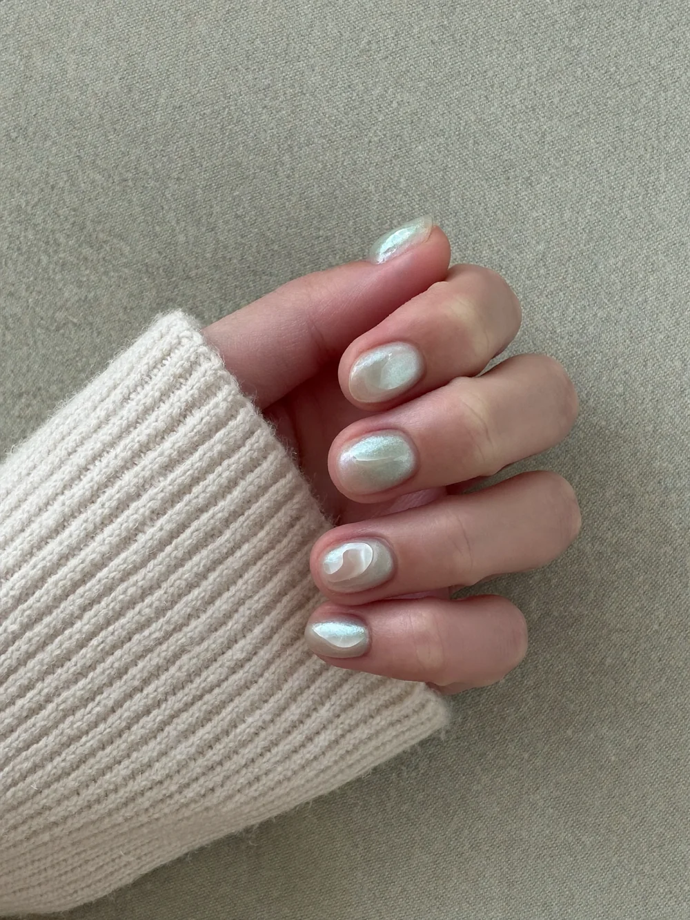 A hand in a cream knit sleeve with short rounded nails in a soft glazed pearl finish