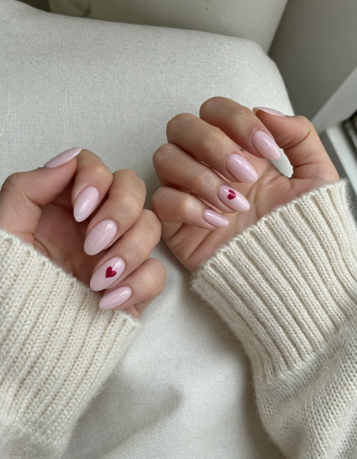Two hands in cream knit sleeves with milky pink almond nails and two tiny burgundy hearts