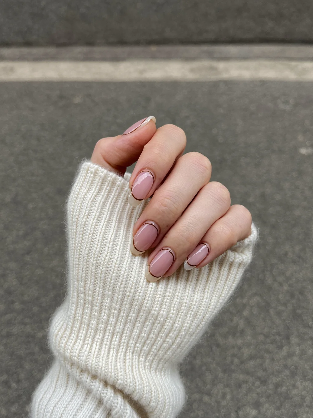 A hand in a cream knit sleeve with sheer pink nails outlined by a thin dark micro French line