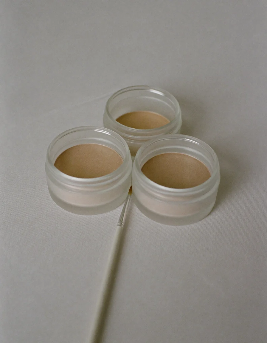 Three small frosted glass jars of pale nude powder beside a slim brush on pale oyster linen