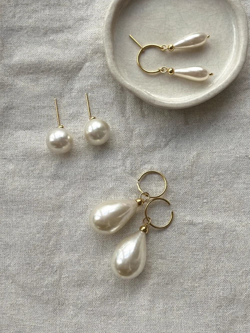 Pearl stud earrings and delicate pearl drop earrings on gold hooks on oyster linen beside a small ceramic dish