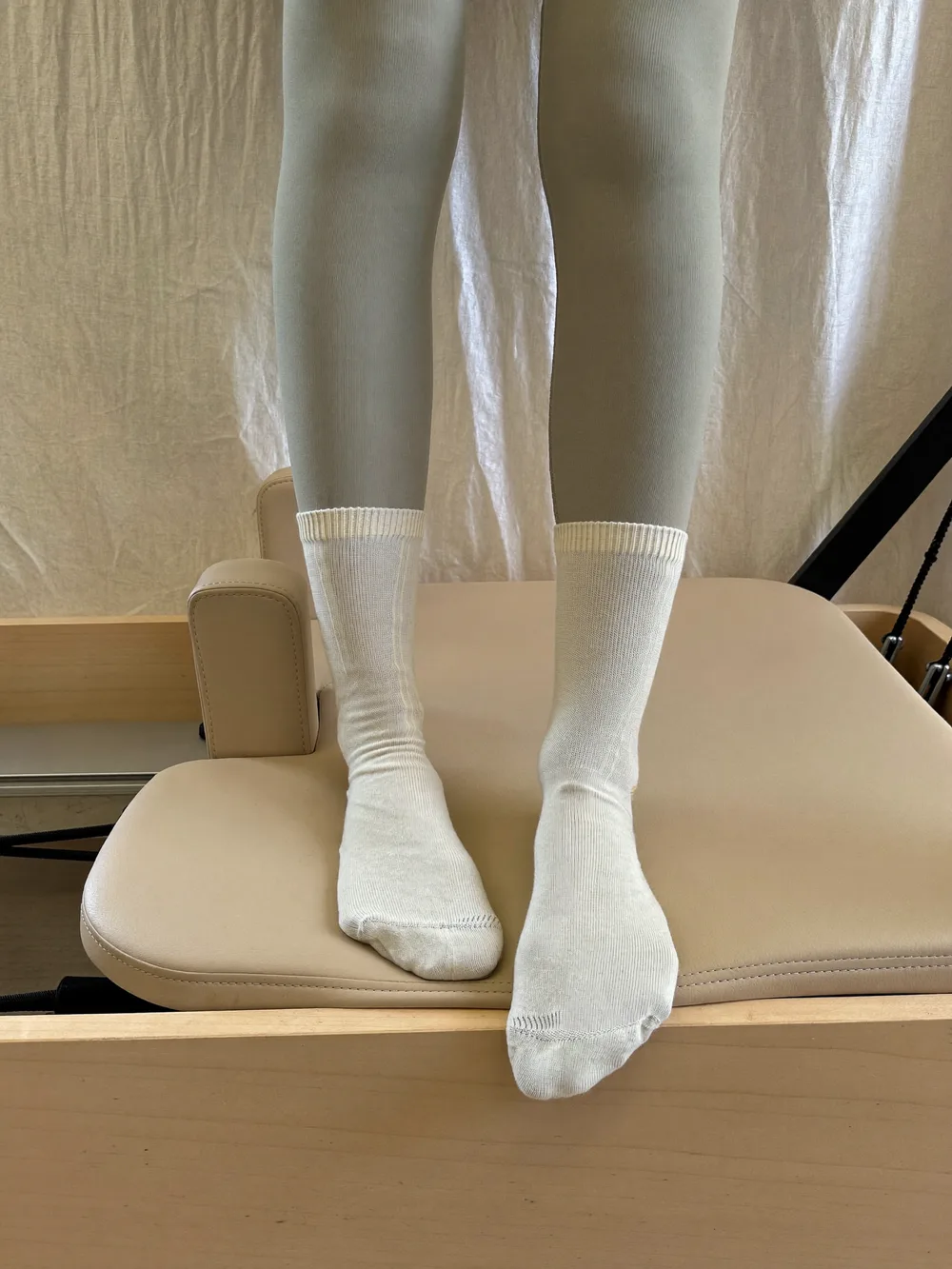 Feet in cream grip socks standing on a Pilates reformer, sage leggings at the ankle