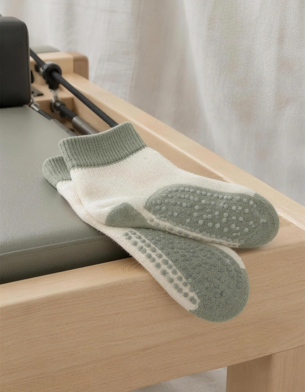 A folded pair of cream and sage grip socks with silicone dots on the wooden edge of a Pilates reformer