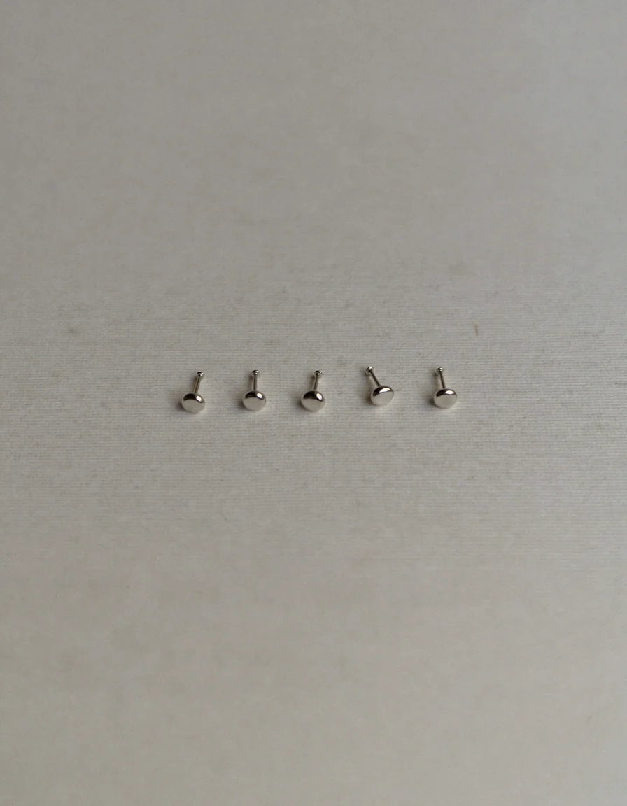 Four tiny plain polished silver stud earrings arranged in a loose row on pale oyster linen