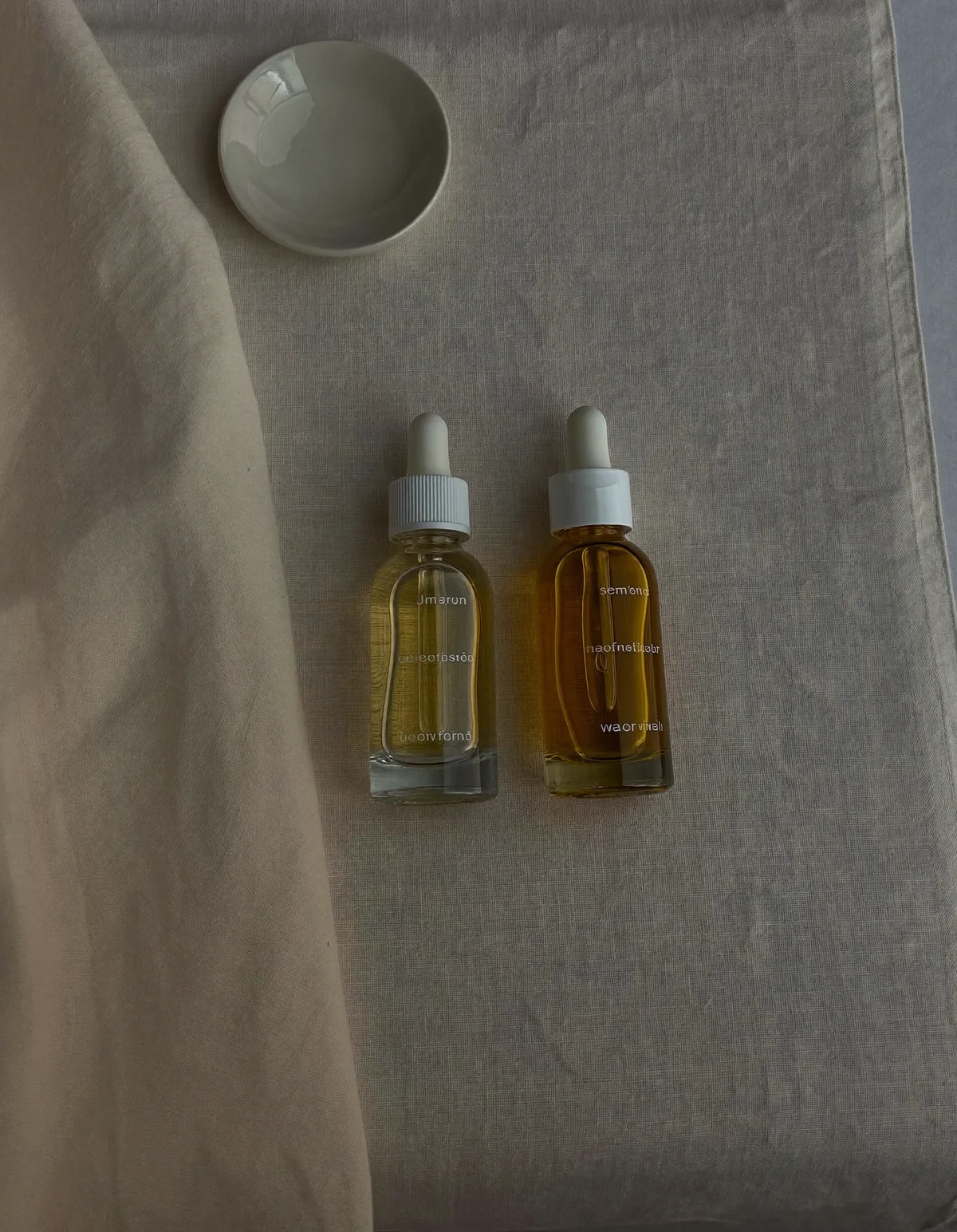 Two plain amber dropper bottles of pale and deeper golden serum on oyster linen beside a small white dish
