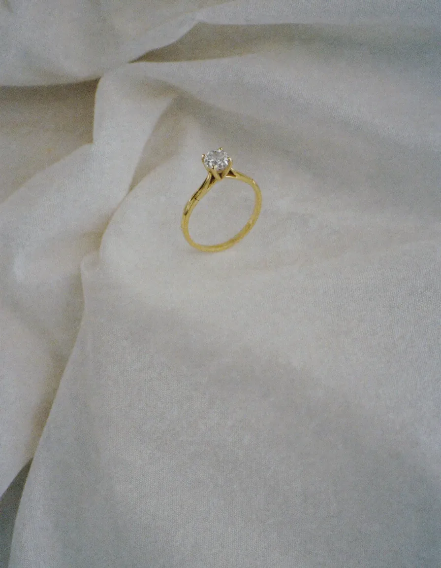 A slim gold solitaire ring shown upright, prongs and band visible, on pale oyster linen